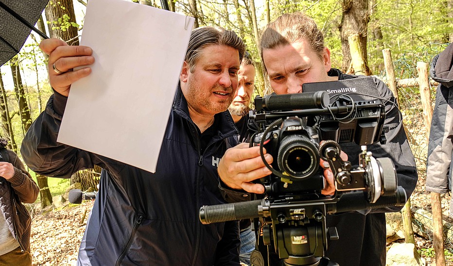 Foto: HSZG Ein Kameramann steht im Wald hinter einer Kamera und stellt diese ein. Ein Assistent hält ein weißes Blatt zum Weißabgleich in der Hand.