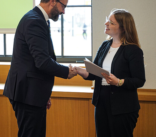 A man in a suit shakes hands with a woman in business attire. Both smile at each other; the woman holds a certificate.