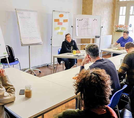 Eine Seminargruppe sitzt im Seminarraum und hört einer Dozierenden zu. Im Hintergrund stehen Aufsteller.