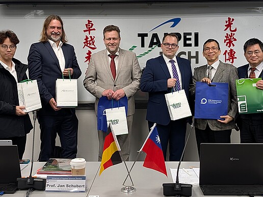 Rektor Alexander Kratzsch und Mitglieder der sächsischen Delegation stehen gemeinsam mit Vertretern der Taipei Tech Universität vor einem Whiteboard und halten Gastgeschenke in den Händen. Auf dem Tisch stehen deutsche und taiwanesische Flaggen.