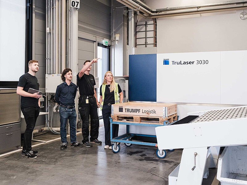 In einer Maschinenhalle stehen drei Männer und eine Frau. Ein Mann zeigt mit ausgestrecktem Arm auf eine Industrieanlage.