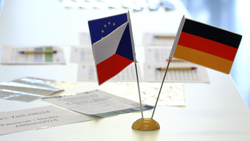 Three miniature national flags of the Czech Republic, Germany and the EU on a white table, with documents scattered next to them.