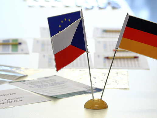 Three miniature national flags of the Czech Republic, Germany and the EU on a white table, with documents scattered next to them.
