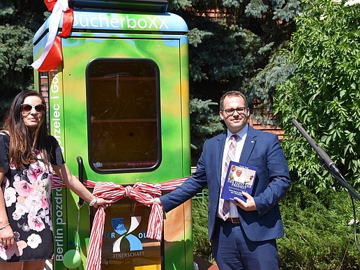 A woman with sunglasses stands on the left and the rector of the HSZG on the right next to a telephone box that has been converted into a bibliobox. They are pulling on a ribbon to symbolically inaugurate the box. They are standing outside, the sun is shining.