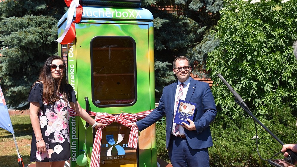 Eine Frau mit Sonnenbrille steht links und der Rektor der HSZG rechts neben einer zur Biblioboxx umgebauten Telefonzelle. Bei ziehen an einem Schleifenband, um die Box symbolisch einzuweihen. Sie stehen im Freien, die Sonne scheint.