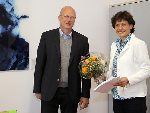 Der Vorsitzende des Hochschulrates und das neue Mitglied Katrin Bartsch stehen im Senatssaal und lächeln in die Kamera. Frau Bartsch hält einen Blumenstrauß in der Hand.
