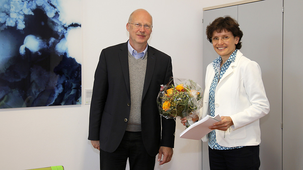 Der Vorsitzende des Hochschulrates und das neue Mitglied Katrin Bartsch stehen im Senatssaal und lächeln in die Kamera. Frau Bartsch hält einen Blumenstrauß in der Hand.