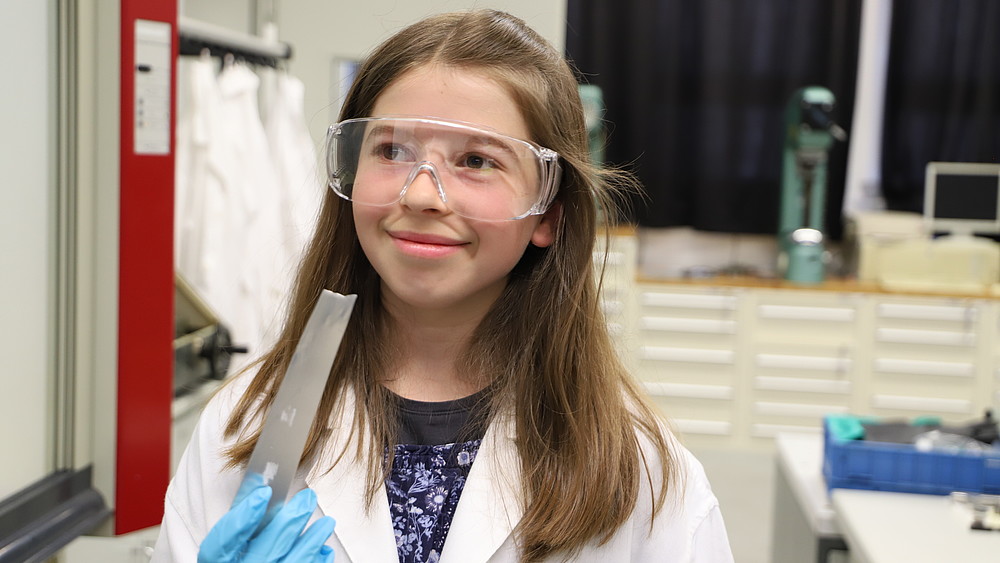 A fifth-grader wears a lab coat and safety goggles and holds a metal workpiece in her hand with a smile.