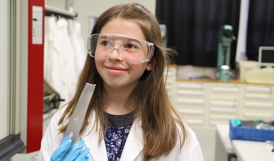 Foto: Antje Pfitzner Eine Fünftklässlerin trägt einen Laborkittel und eine Schutzbrille und hält lächelnd ein Metallwerkstück in der Hand.