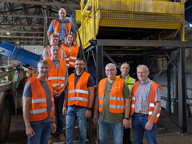 Eine Gruppe von neun Personen steht in einer Industriehalle vor einer großen gelben Sortier- oder Recyclinganlage. Alle tragen orange Sicherheitswesten und blicken in die Kamera.
