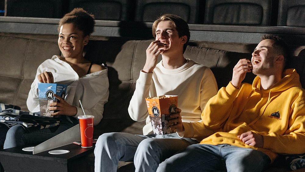 Three young moviegoers sit in the cinema eating popcorn and laughing.