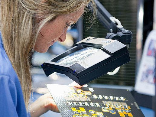 A blonde woman bends over a circuit board with an electronic circuit.