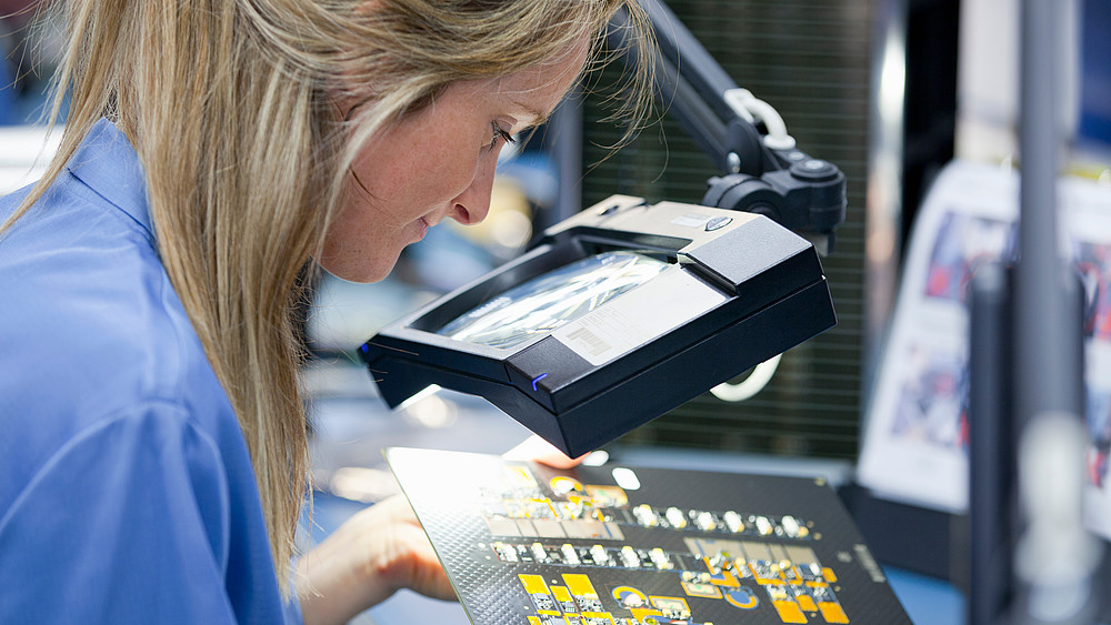 A blonde woman bends over a circuit board with an electronic circuit.