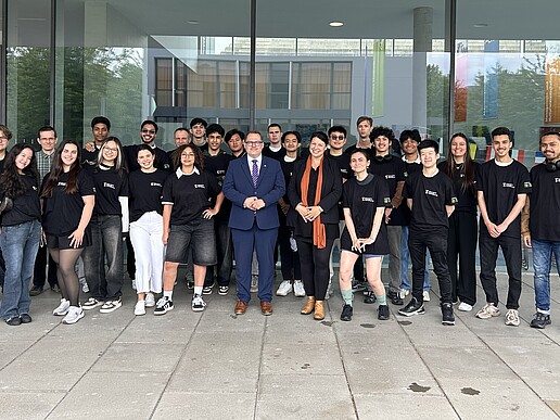 Eine Gruppe ausländischer Studierender und der Rektor stehen vor der Glasfassade von Haus Z vier und lächeln in die Kamera.