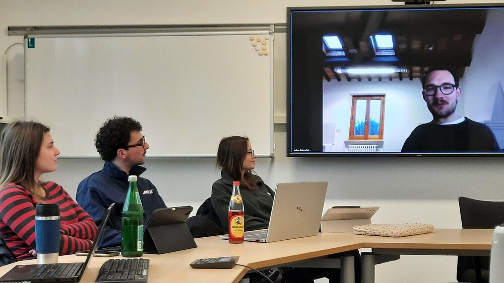 Students sit at a table in the seminar room and look at a screen on which the author Lukas Rietzschel is connected.