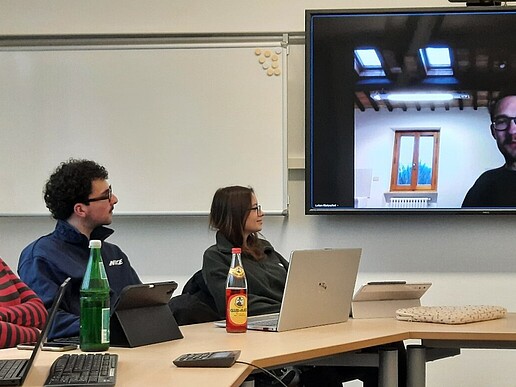 Students sit at a table in the seminar room and look at a screen on which the author Lukas Rietzschel is connected.