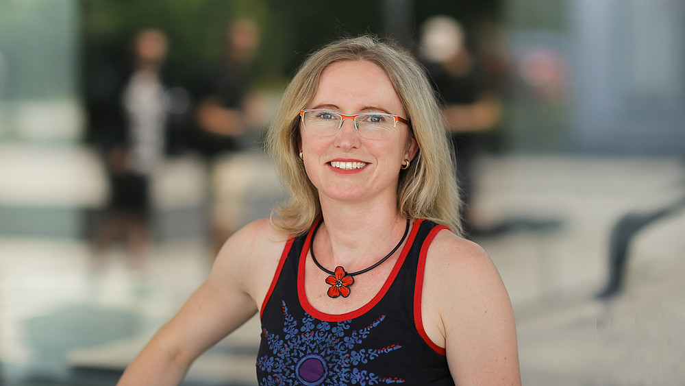 Dr. rer. nat. Kateřina Barková, a blonde woman with glasses in a summery top with a red flower necklace, smiles at the camera.