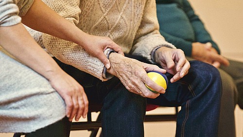 Ein älterer Herr sitzt mit einem gelben Ball in der Hand auf einem Stuhl. Neben ihn sitzt eine jüngere Frau. Sie hält und unterstützt seinen Arm.