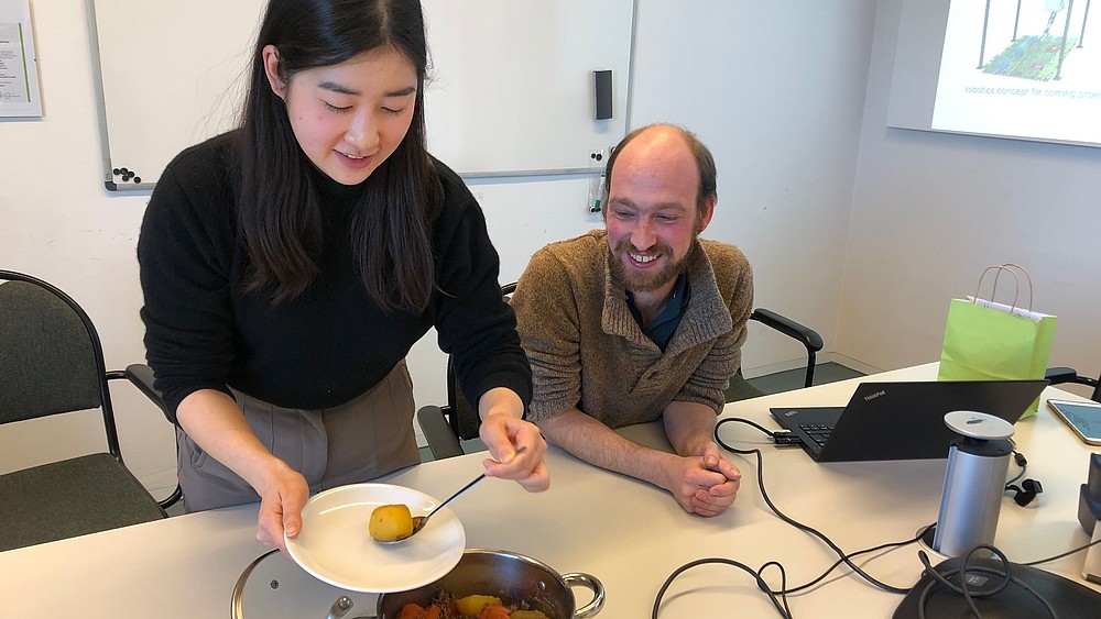 A young Japanese woman fills a plate with stew. A staff member sits next to her at the table, smiling.