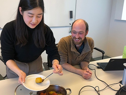 A young Japanese woman fills a plate with stew. A staff member sits next to her at the table, smiling.