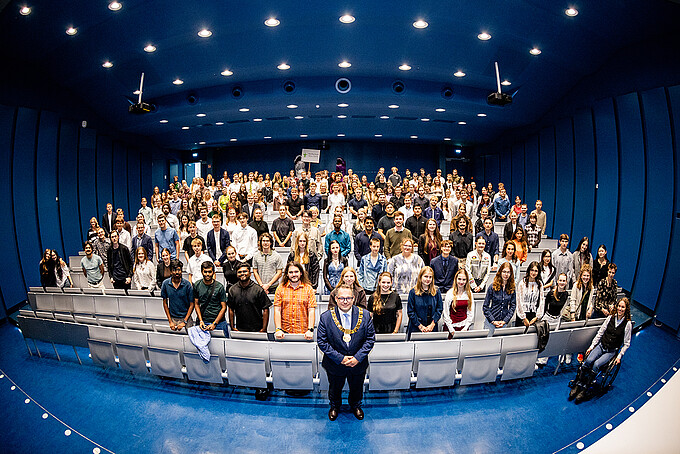 A large group of first-year students in the Audimax on the Zittau campus; in the front is Rector Prof. Dr.-Ing. Alexander Kratzsch; among the students, two people dressed as dinosaurs can also be seen.