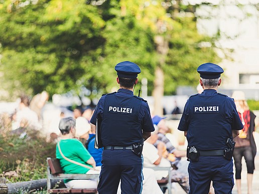 Two police officers stand in a park with their backs to the camera, facing forward.