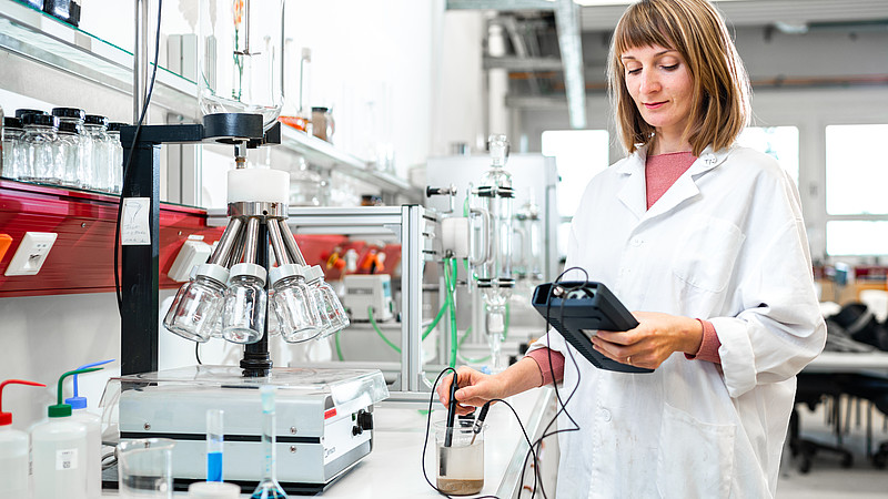 Eine Laborantin im weißen Kittel arbeitet im Labor und hält ein Messgerät in der Hand.