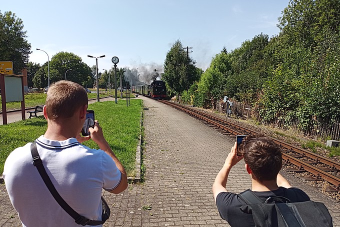 Two young men stand with their backs to the camera and take pictures of the arriving train with their cell phones