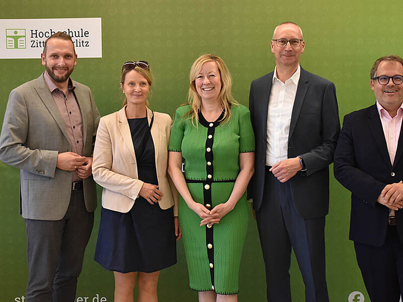 A delegation from the HSZG and the city of Bautzen stands in front of the green backdrop and smiles for the camera.
