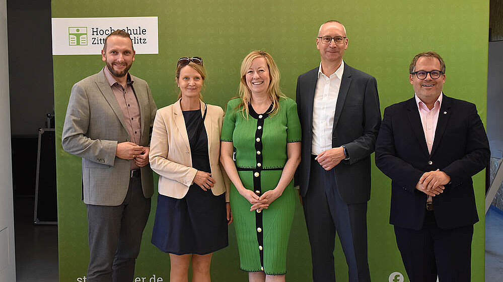 A delegation from the HSZG and the city of Bautzen stands in front of the green backdrop and smiles for the camera.