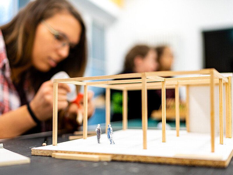 A student uses craft glue to build a model house out of wooden sticks. The model stands on the table in front of her.