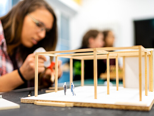 Eine Studentin benutzt Bastelkleber, um ein Hausmodell aus Holzstäben zu bauen. Das Modell steht vor ihr auf dem Tisch.