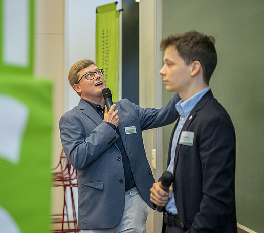 Two young scientists explain their project on a green board.