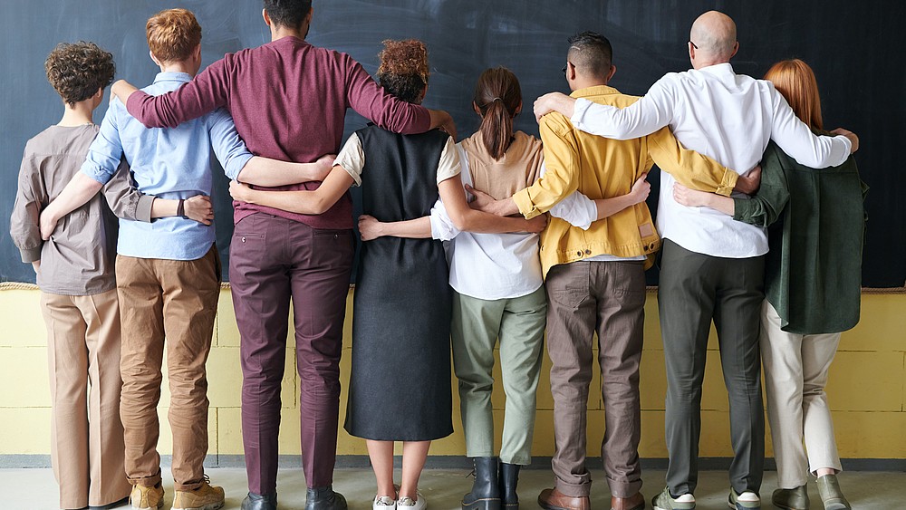 A number of young people of different ethnic backgrounds stand in a row, their backs turned to the camera, facing a blackboard. They place their hands and arms on the person standing next to them.