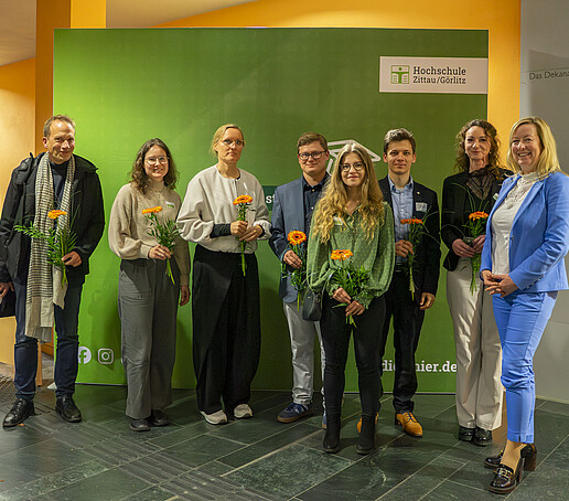 Speakers and the Vice President stand smiling next to each other in front of a green HSZG press wall for a group photo.