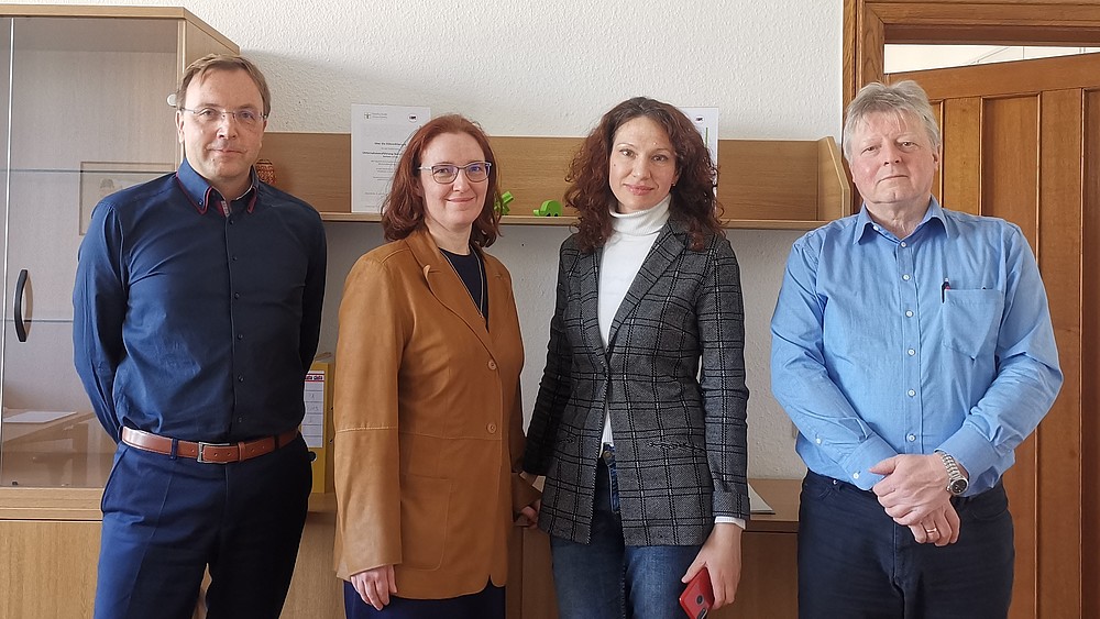 Four people stand next to each other in a room and look into the camera.