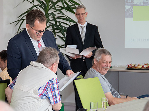 Ältere ehemalige Studierende sitzen an einer Tafel im Senatssaal. Der Rektor und Prof. Bellair stehen im Hintergrund und unterhalten sich mit ihnen.