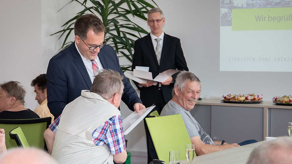 Ältere ehemalige Studierende sitzen an einer Tafel im Senatssaal. Der Rektor und Prof. Bellair stehen im Hintergrund und unterhalten sich mit ihnen.