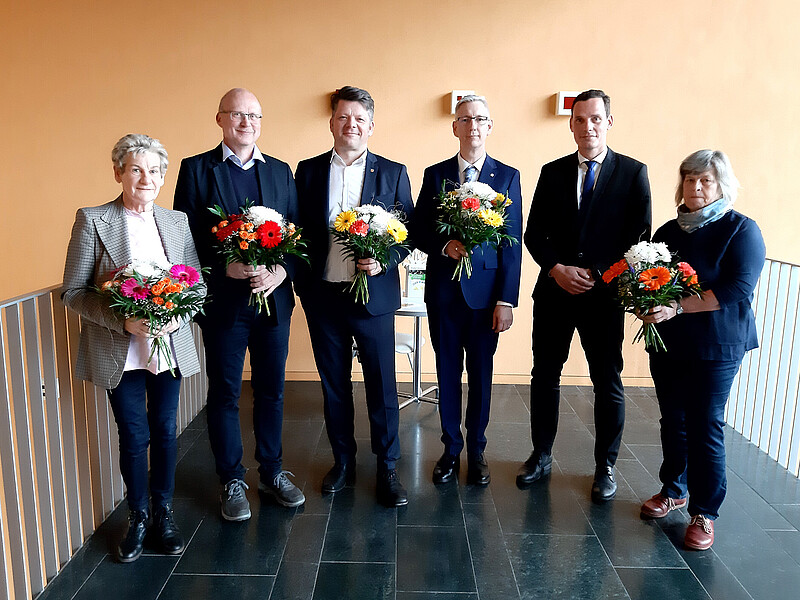 Sechs Personen des Hochschulrates stehen im Flur, halten Blumensträuße und lächeln in die Kamera.