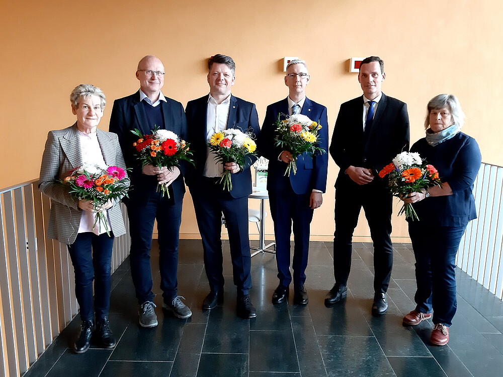 Six members of the University Council stand in the hallway, holding bouquets of flowers and smiling at the camera.