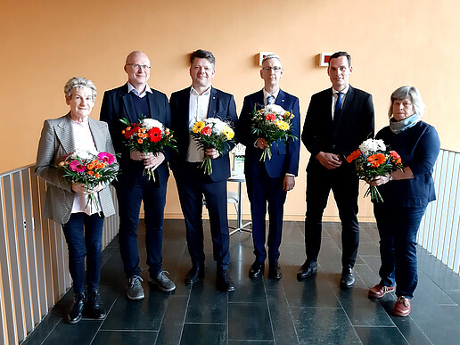 Six members of the University Council stand in the hallway, holding bouquets of flowers and smiling at the camera.