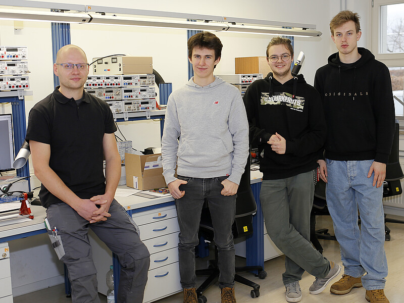 In the training workshop, the three new trainees are standing in front of a table, with the trainer leaning against the table to their left.
