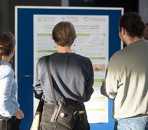 Three people stand in front of a poster wall and look at a poster with research results on it.