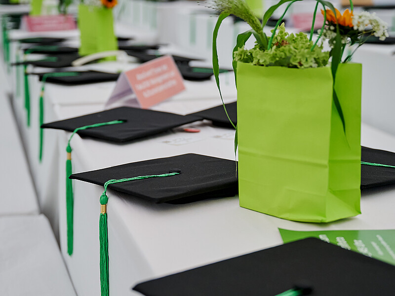 Auf einem mit weißer Tischdecke bedeckten Tisch liegen Absolventenhüte und in grüne Papiertüten gesteckte Blumensträuße.