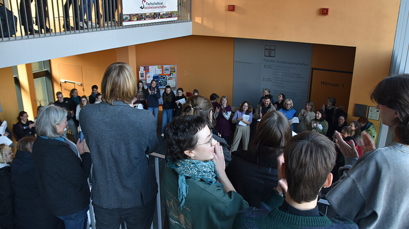 Several people are standing in the stairwell of a campus building, holding sheet music and singing.