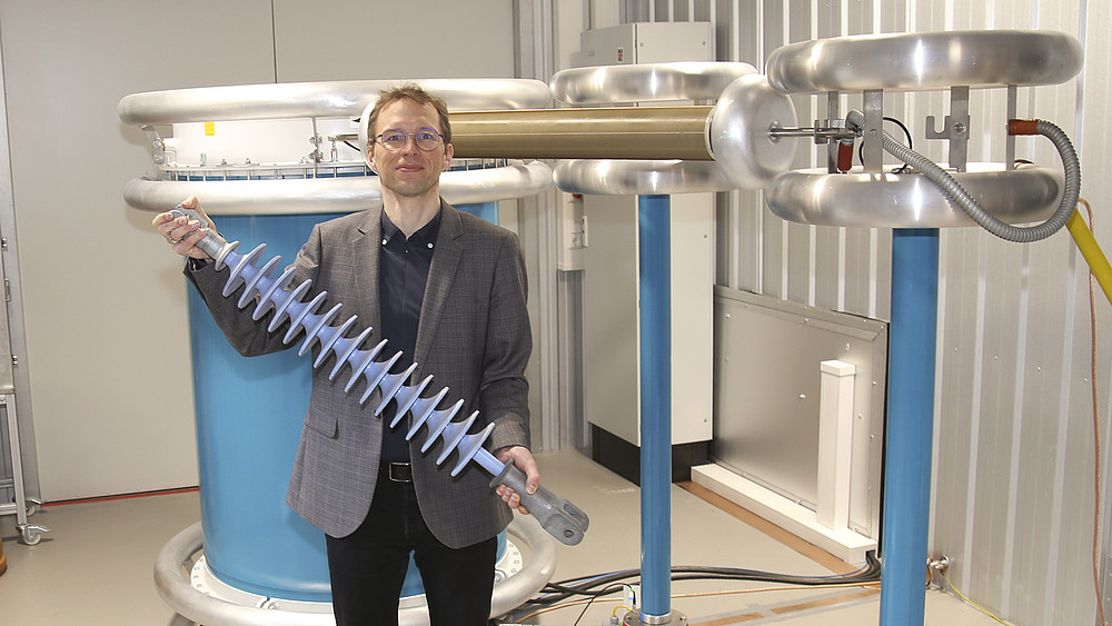 A professor stands in front of a voltage system and holds a component in his hand.