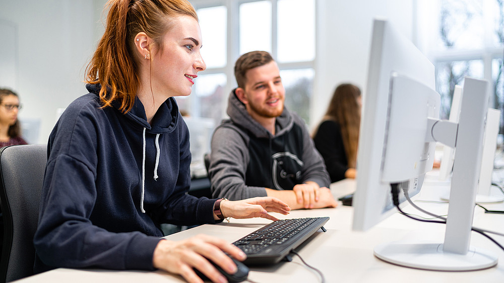 Eine Studentin und ein Student sitzen im Computerkabinett vor einem PC-Bildschirm und wenden diesem ihren Blick zu.