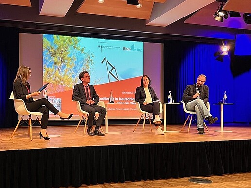 Vier Menschen sitzen auf einem Podium und diskutieren. Im Hintergrund leuchtet eine Projektion.