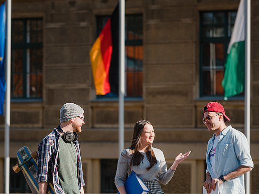 Die europäische, deutsche und sächsische Flagge fliegen vor einem Hochschulgebäude in Görlitz. Davor sind drei Studierende im Gespräch.