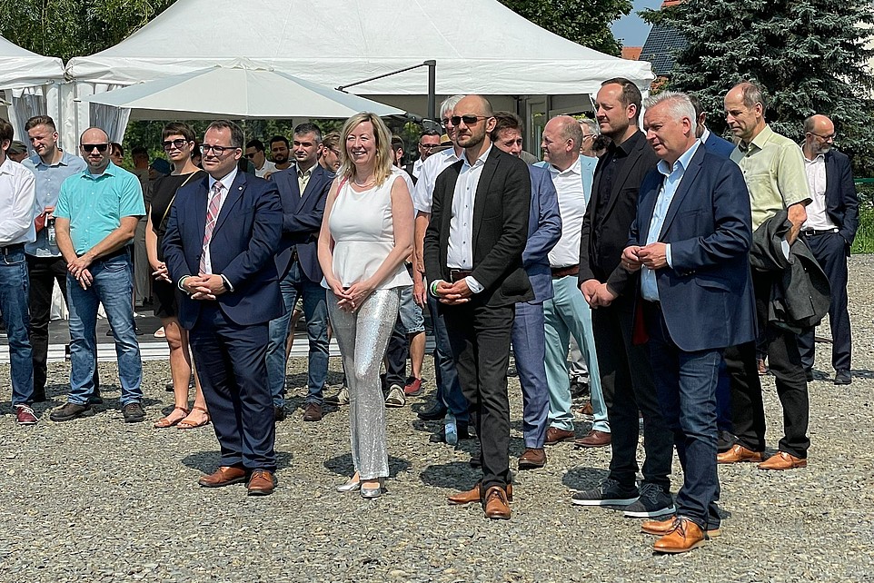 The university management and guests stand outside in front of a pavilion and listen to someone who is not in the picture.